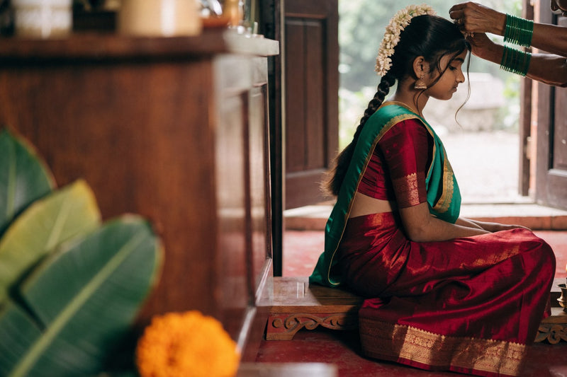 A girl seated in a crimson and green silk langa voni at her half saree function while an elder places jasmine in her braid