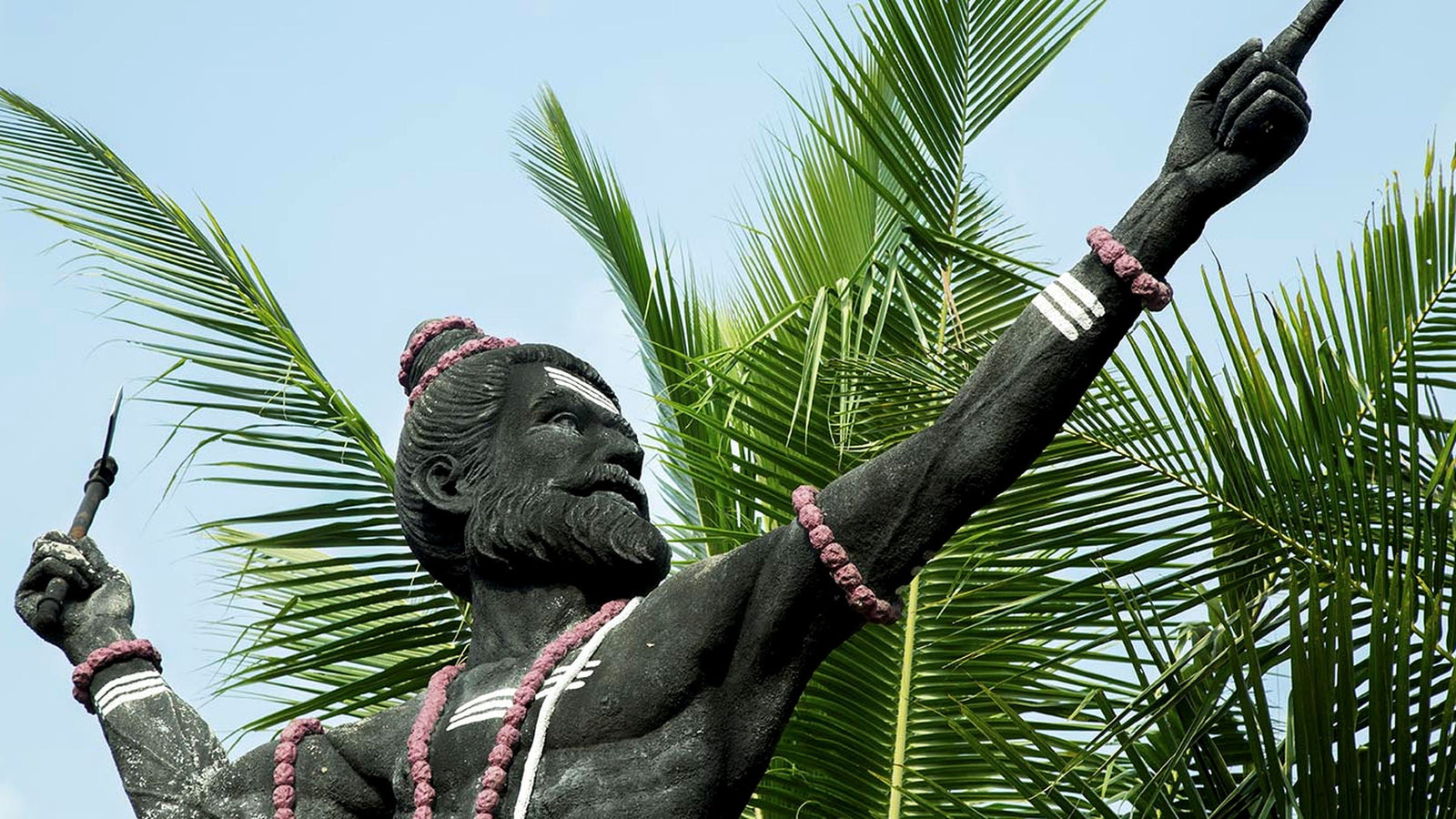 A dark stone statue of Parashurama with matted hair, rudraksha beads and an axe, raised against palm fronds