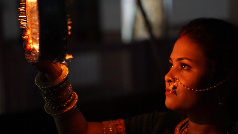 A woman in festive jewellery raises a decorated sieve towards the night sky on Karwa Chauth.